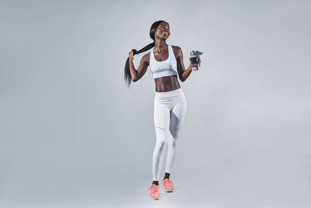 Happy young African woman in sports clothing holding bottle with water and adjusting hair - Foto, Imagem