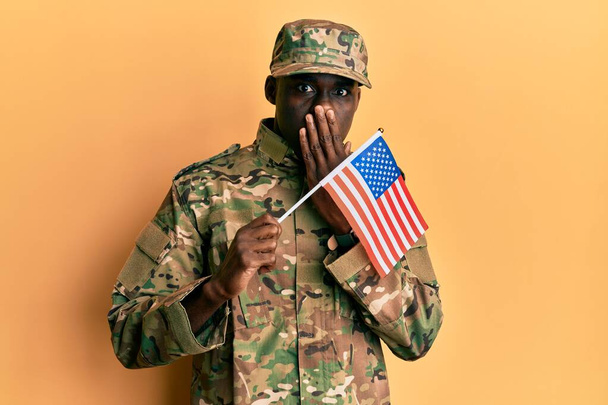 Молодой африканский американец в армейской форме с американским флагом, прикрывающим рот рукой, шокирован и боится ошибиться. Удивительное выражение  - Фото, изображение