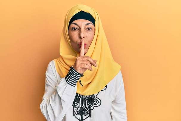 Middelbare leeftijd Spaanse vrouw draagt traditionele islamitische hijab sjaal vragen om stil te zijn met de vinger op de lippen. stilte en geheim concept.  - Foto, afbeelding