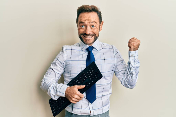 Middle age businessman holding keyboard screaming proud, celebrating victory and success very excited with raised arm  - Photo, Image