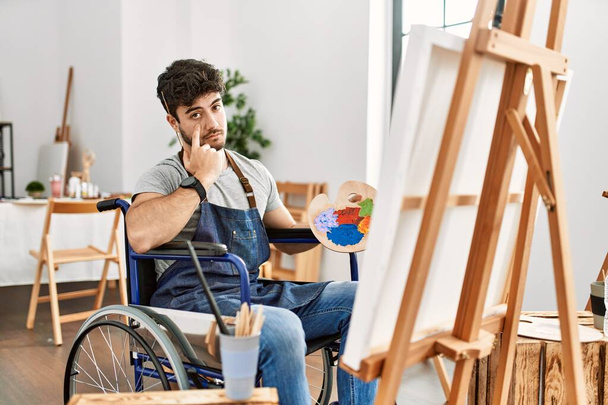 Joven hombre hispano sentado en una silla de ruedas pintando en un estudio de arte apuntando al ojo observándote gesto, expresión sospechosa  - Foto, imagen