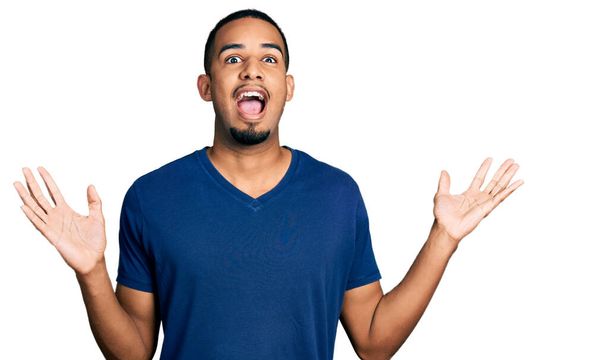 Joven hombre afroamericano vistiendo camiseta casual celebrando loco y sorprendido por el éxito con los brazos levantados y los ojos abiertos gritando emocionado. concepto ganador  - Foto, imagen