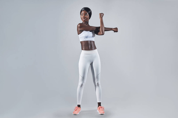 Full length of beautiful young African woman in sports clothing stretching against gray background - Fotografie, Obrázek