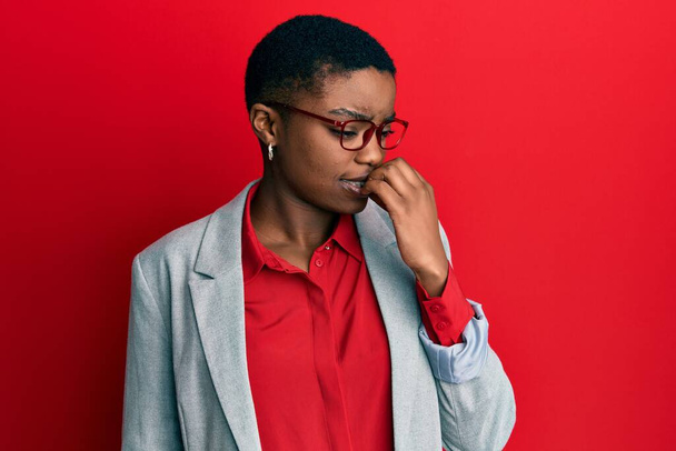 Młoda Afroamerykanka w biznesowej kurtce i okularach wyglądająca na zestresowaną i zdenerwowaną z rękami obgryzającymi paznokcie. niepokój.  - Zdjęcie, obraz