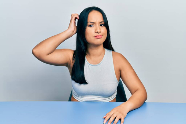 Schöne hispanische Frau mit Nasenpiercing, die auf dem Tisch sitzt, verwirrt und wundert sich über die Frage. Unsicher im Zweifel, mit der Hand auf dem Kopf denkend. Nachdenkliches Konzept.  - Foto, Bild