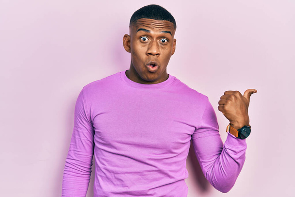 Junger schwarzer Mann in lässigem rosafarbenem Pullover überrascht mit erhobenem Zeigefinger zur Seite, offener Mund erstaunter Ausdruck.  - Foto, Bild