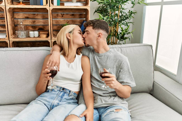 Jovem casal caucasiano beijando e brindando com vinho tinto em casa. - Foto, Imagem
