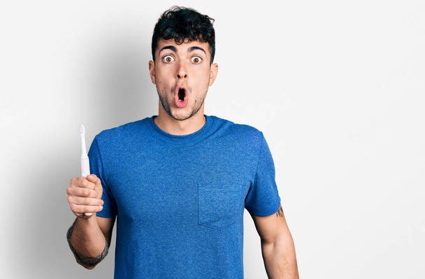 Jonge Spaanse man houdt elektrische tandenborstel bang en verbaasd met open mond voor verrassing, ongeloof gezicht  - Foto, afbeelding