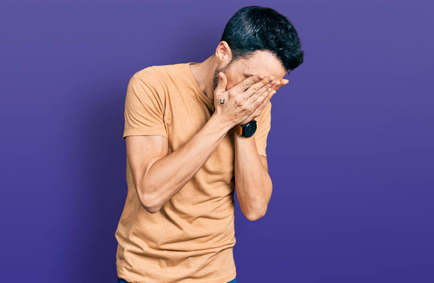 Hispánec s vousy, oblečený do neformální trička se smutným výrazem, zakrývající obličej rukama, zatímco brečí. deprese koncept.  - Fotografie, Obrázek