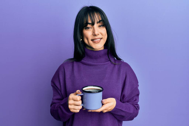 Giovane donna bruna con frangia che beve una tazza di caffè dall'aspetto positivo e felice e sorride con un sorriso fiducioso mostrando i denti  - Foto, immagini