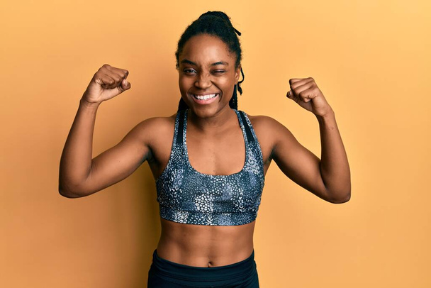 African american woman with braided hair wearing sportswear showing arm muscles winking looking at the camera with sexy expression, cheerful and happy face.  - Фото, изображение