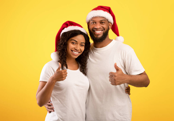 Gelukkig Nieuwjaar. Vrolijke zwarte echtgenoten in Santa hoeden tonen duimen omhoog en glimlachen om de camera op gele achtergrond - Foto, afbeelding