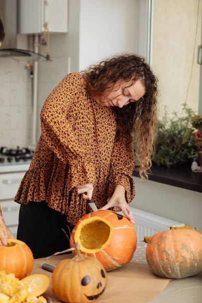 Close up of young woman carving Jack O Lattern from ripe orange pumpkin with knife on her wooden kitchen table. Female preparing all hallows eve Halloween party decorations. Background, copy space. - Photo, Image