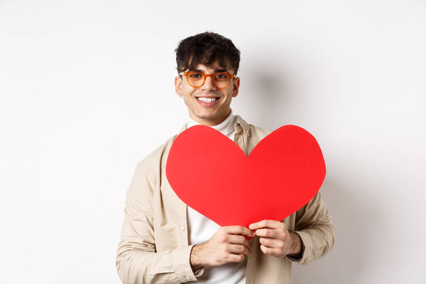 Guapo chico hipster con gafas de sol y pendientes, esperando el verdadero amor en el día de San Valentín, sosteniendo un gran corazón rojo y sonriendo, de pie sobre fondo blanco - Foto, imagen