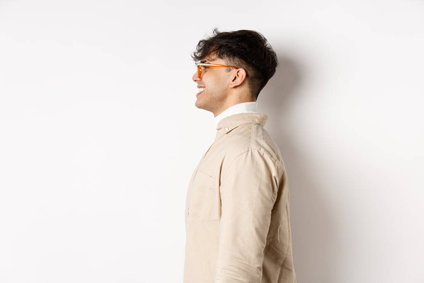 Profile of handsome caucasian man in eyewear, looking at empty space left with cheerful smile, standing on white background - Fotografie, Obrázek