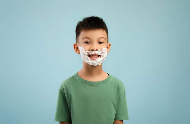 Petit garçon asiatique avec mousse à raser sur le visage posant sur fond bleu - Photo, image