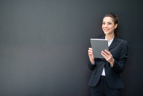 Χαμογελώντας νεαρή επιχειρηματίας χρησιμοποιώντας το tablet της μπροστά από ένα μαύρο τοίχο παρόμοια με έναν πίνακα - Φωτογραφία, εικόνα