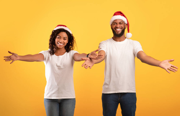 Komm her, lass mich dich umarmen. Freundliches afrikanisch-amerikanisches Paar in Weihnachtsmützen, ausgestreckte Hände nach vorne, gelber Hintergrund - Foto, Bild