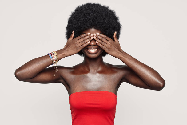 Beautiful young African woman covering eyes with hands and smiling while standing against gray background - Foto, Imagem