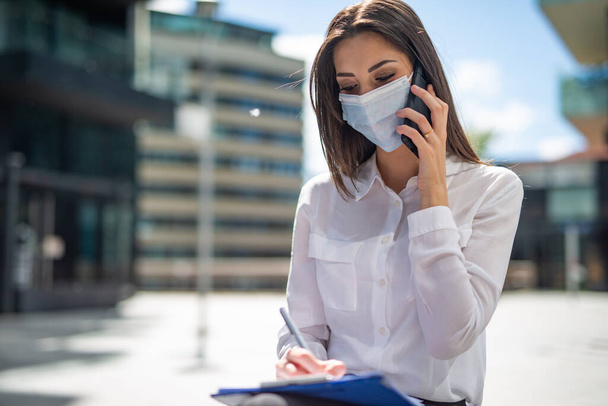 Masked young woman talking on the phone while writing notes, covid and coronavirus concept - Foto, afbeelding