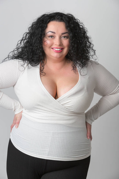 Plus size woman with friendly toothy smile. Oversized model with deep cleavage, big breast and long curly dark hair posing in studio. Overweight female with white teeth and red lipstick - Photo, Image