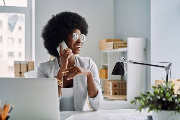 Krásná mladá africká žena mluví na mobilním telefonu a usmívá se, zatímco sedí na svém pracovišti v kanceláři - Fotografie, Obrázek