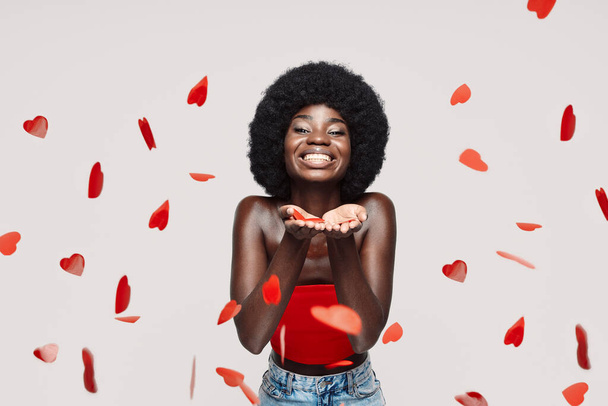 Beautiful young African woman looking at camera and blowing confetti while standing against gray background - Fotografie, Obrázek