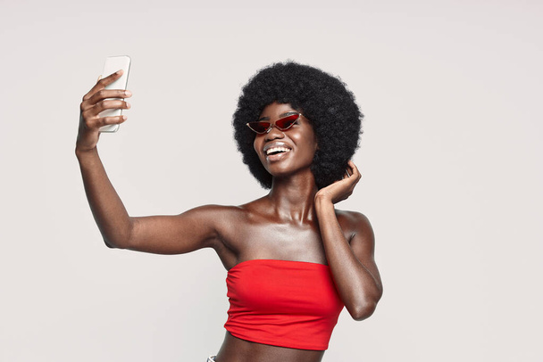 Beautiful young African woman making selfie by smart phone and smiling while standing against gray background - Foto, Imagem