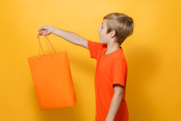 the boy stretched out his hand to the side in which he holds an orange shopping bag - Foto, afbeelding