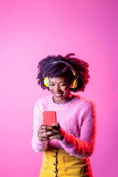 Isolierte schwarze junge Frau mit Smartphone beim Videoanruf mit drahtlosen Kopfhörern auf Werbe-Copyspace-Hintergrund - Foto, Bild