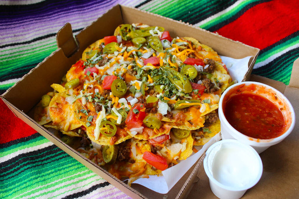 Neem een doos met nacho 's belegd met rundvlees, jalapenos, tomaten, uien en kaas, met salsa en zure room aan de zijkant om te dompelen - Foto, afbeelding