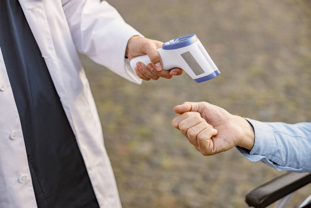 Nahaufnahme eines medizinischen Infrarot-Thermometers in der Hand eines Arztes, der die Temperatur eines behinderten Patienten im Rollstuhl während des Covid19-Ausbruchs misst - Foto, Bild
