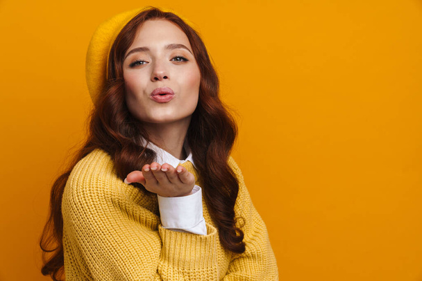 Happy young woman with long red hair in sweater and beret standing over yellow wall background, sending love kiss - 写真・画像