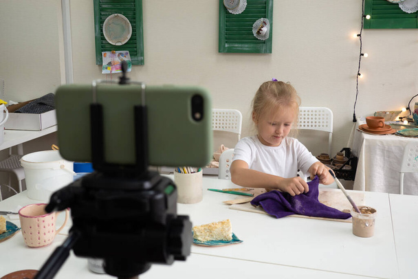 Girl vlogger recording video with modelling clay and speaking for smartphone camera. Kid blogger in a ceramic studio creating interesting content for children.  - Foto, afbeelding