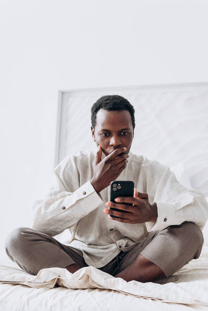 Sonriente hombre negro olas mano en videocall sentado en la cama - Foto, Imagen