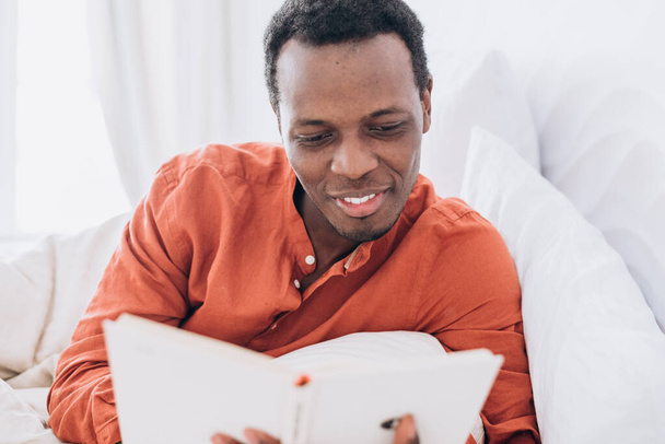 Hombre negro en pijama lee libro acostado en cama cómoda - Foto, imagen