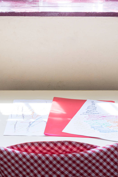 Photograph of some drawings and a red folder of a child on a table in a classroom of a nursery school.The photo is taken in vertical format. - Photo, Image
