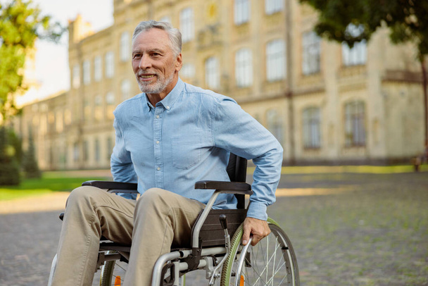 Sonriendo anciano hombre recuperado en silla de ruedas mirando hacia otro lado mientras pasa tiempo solo en el parque cerca del hospital - Foto, Imagen