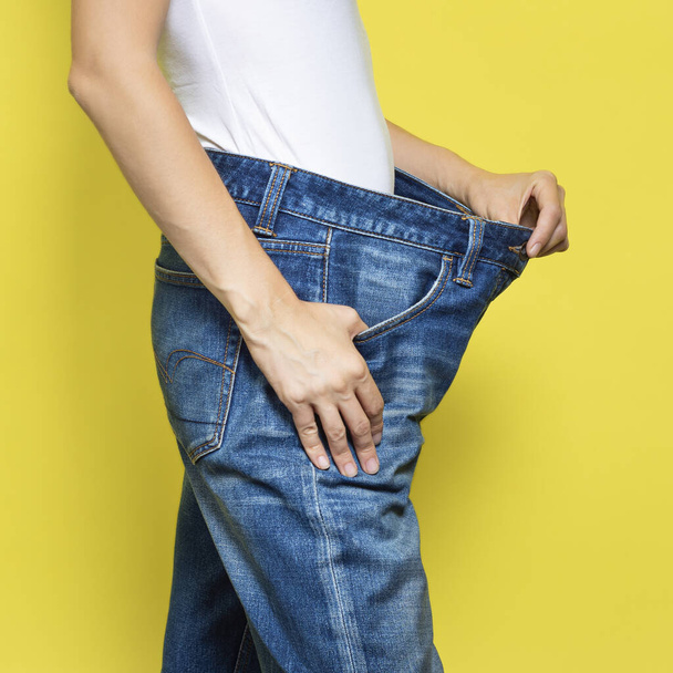 Das Konzept der Ernährung, richtige Ernährung, Gewichtsverlust. Schlanke Frau mit lockeren Jeans und ihrem Abnehmen. Frau weißes T-Shirt und Oversize-Jeans auf gelbem Hintergrund - Foto, Bild