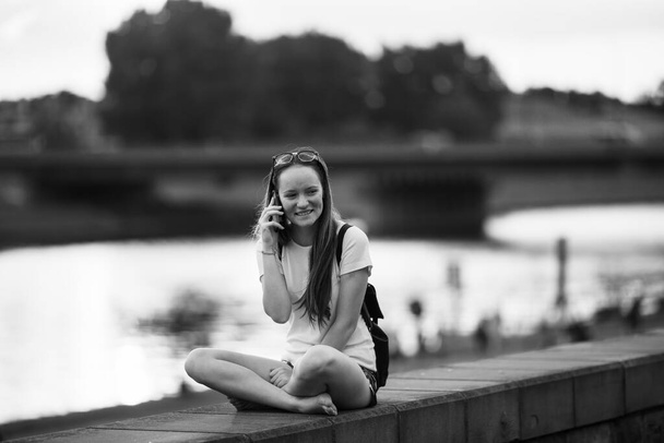 Μια έφηβη που κάθεται στην προκυμαία και μιλάει σε ένα κινητό τηλέφωνο... ασπρόμαυρη φωτογραφία. - Φωτογραφία, εικόνα