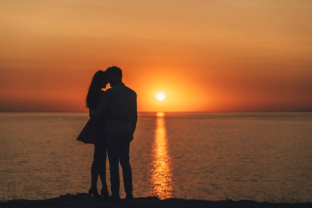 Het jonge stel dat aan de kust van de zee staat op de schilderachtige achtergrond van de zonsondergang - Foto, afbeelding
