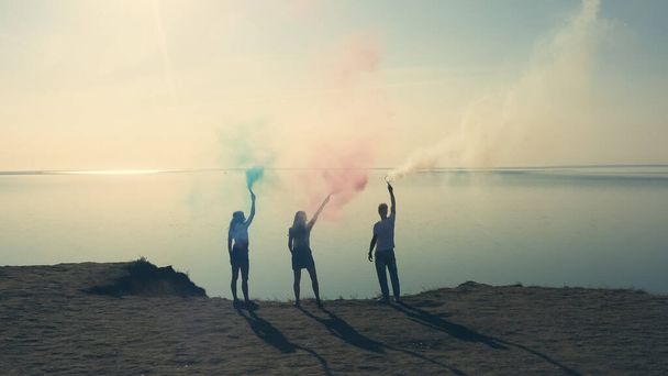 Les trois personnes avec des bâtons de fumée de couleur se tiennent sur le rivage de la mer - Photo, image