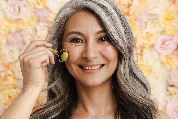 Smiling mature woman massaging face with jade roller standing over floral background - Φωτογραφία, εικόνα