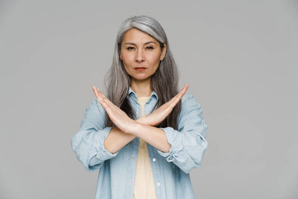 Mature white-haired woman looking at camera and showing stop gesture isolated over gray wall - Foto, immagini