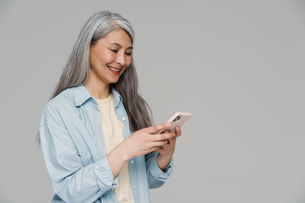 Mature white-haired woman smiling and using mobile phone isolated over gray wall - Foto, afbeelding
