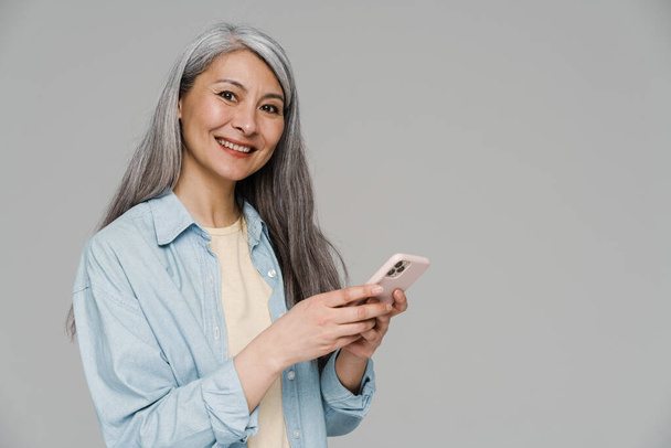 volwassen vrouw met wit haar glimlachen en met behulp van mobiele telefoon geïsoleerd over grijze muur - Foto, afbeelding
