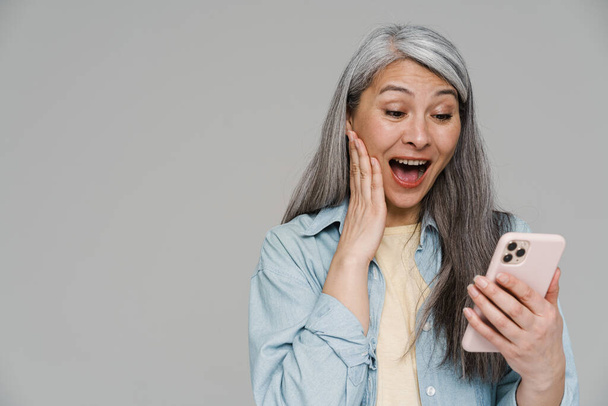 Mature excited woman screaming and using mobile phone isolated over gray wall - Valokuva, kuva