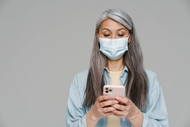 Mature white-haired woman in face mask using mobile phone isolated over gray wall - Foto, afbeelding