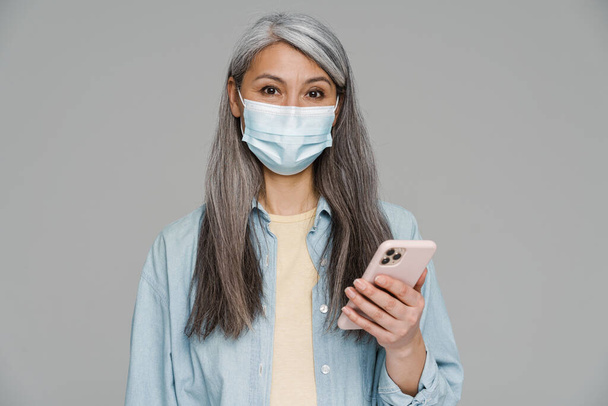 Mature white-haired woman in face mask using mobile phone isolated over gray wall - Foto, afbeelding