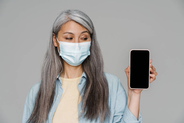 Mature white-haired woman in face mask showing mobile phone isolated over gray wall - Foto, afbeelding